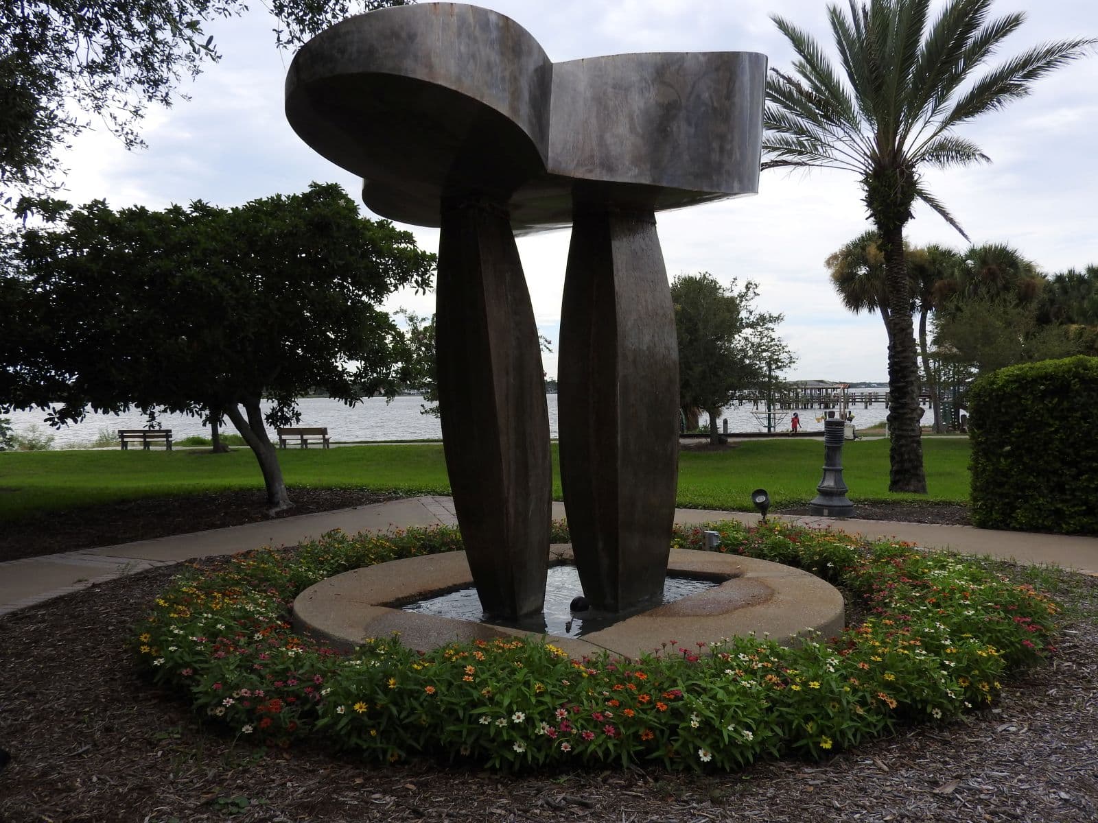 Modern fountain sculpture surrounded by flowers with the Halifax River beyond.